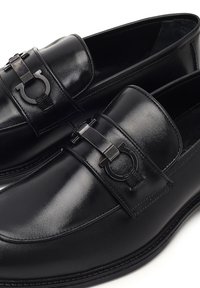 Schwarze Lederloafer mit abgerundetem Zehenbereich, dekorativem Riemen mit metallic Akzent und glatter, polierter Oberfläche.