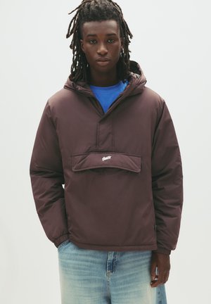 Jachetă pulover închisă la culoare, cu glugă, cu buzunar frontal și manșete elastice; purtată peste o cămașă albastră; textura netedă, design casual, branding minimal.