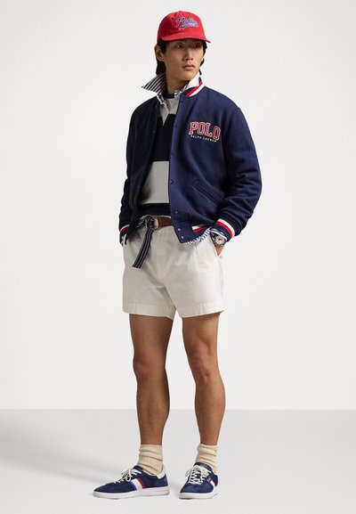 Casaco varsity azul-marinho com detalhes em vermelho e branco, gola listrada, polo cinza, calções beiges e ténis azuis com risca vermelha.