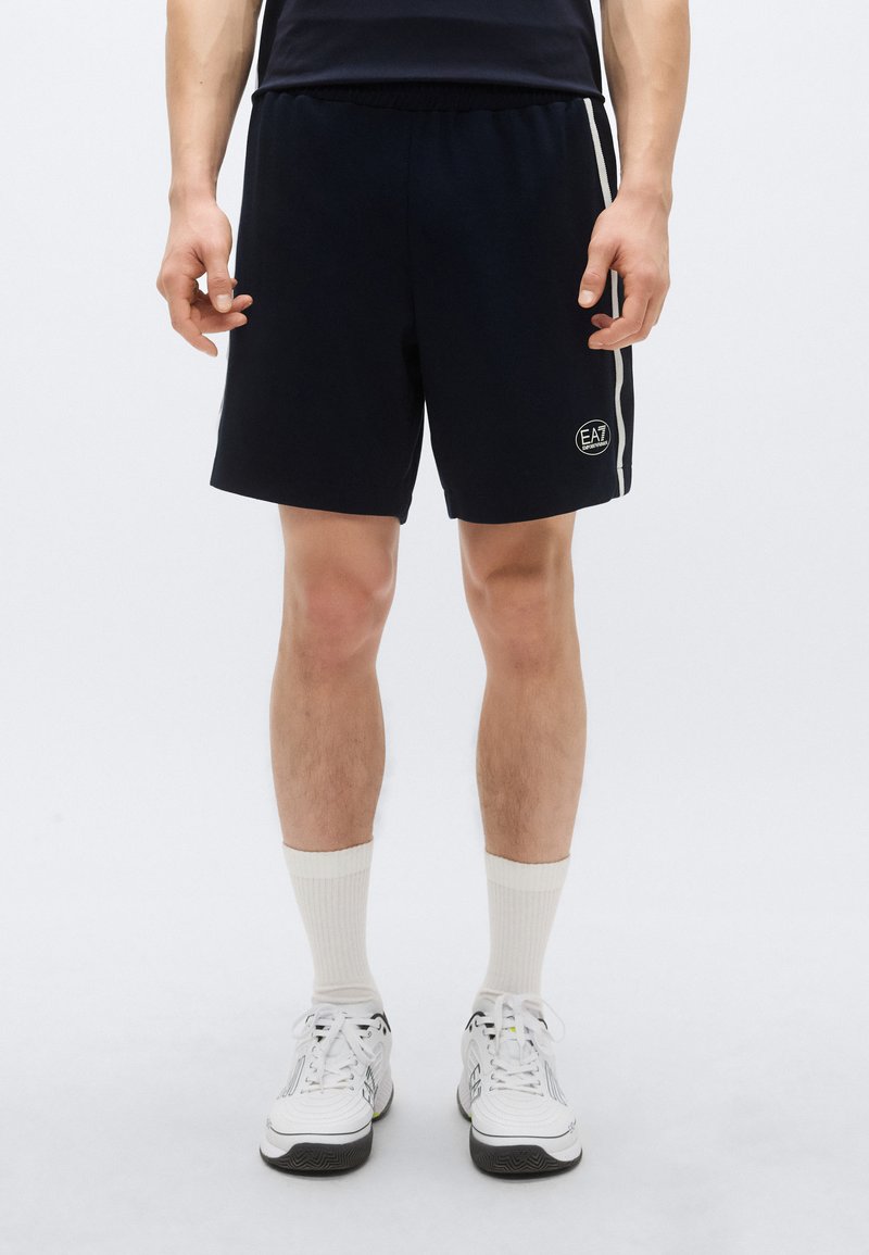 Tmavomodré športové šortky s elastickým pásom, s bielymi bočnými pruhmi a nášivkou s logom. Nosené s bielymi ponožkami a teniskami.