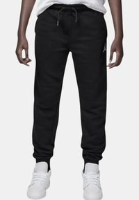 Pantaloni della tuta neri con vita regolabile, gambe affusolate e logo ricamato sulla coscia sinistra, abbinati a sneakers bianche.