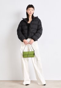 Melnās pūkainās jakas ar kapuci, kurai ir quiltēta dizaina, kombinēta ar vaļīgām baltām kravas biksēm un mazu zaļu rokassomu ar siksnu.