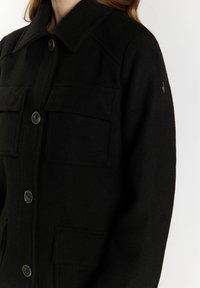 Zwarte wolmix jas met een puntige kraag, twee borstzakken, knopen aan de voorkant en een logo detail aan de zijkant. Gladde textuur en gestructureerde vorm.