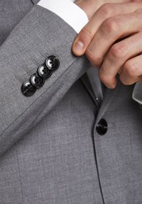 Graue Anzugjacke aus strukturiertem Stoff. Vier schwarze Knöpfe am Ärmel, kombiniert mit einem weißen Hemdärmel darunter. Einfaches, klares Design.
