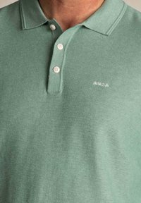 Polo shirt in lichtgroen, gemaakt van gestructureerde stof. Heeft een knoopsluiting met drie knopen en een wit geborduurd logo op de borst.