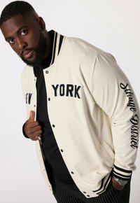 Krämfärgad collegejacka med svart randig ribbad krage och muddar. "NEW YORK" i stora svarta bokstäver på framsidan, dekorationstext på ärmen.