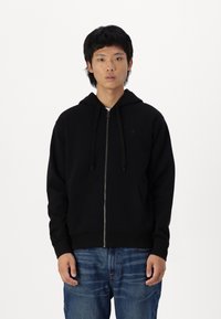 Zwarte rits hoodie met een voorkant zak, verstelbare koordjes en geribde manchetten, gemaakt van een zachte, gestructureerde stof. Draagt met blauwe jeans.