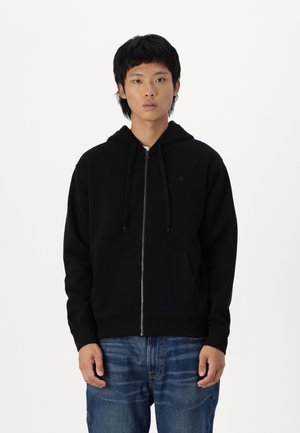 Sudadera con cremallera negra, con un bolsillo frontal, cordones ajustables y puños acanalados, hecha de una tela suave y texturizada. Llevada con jeans azules.