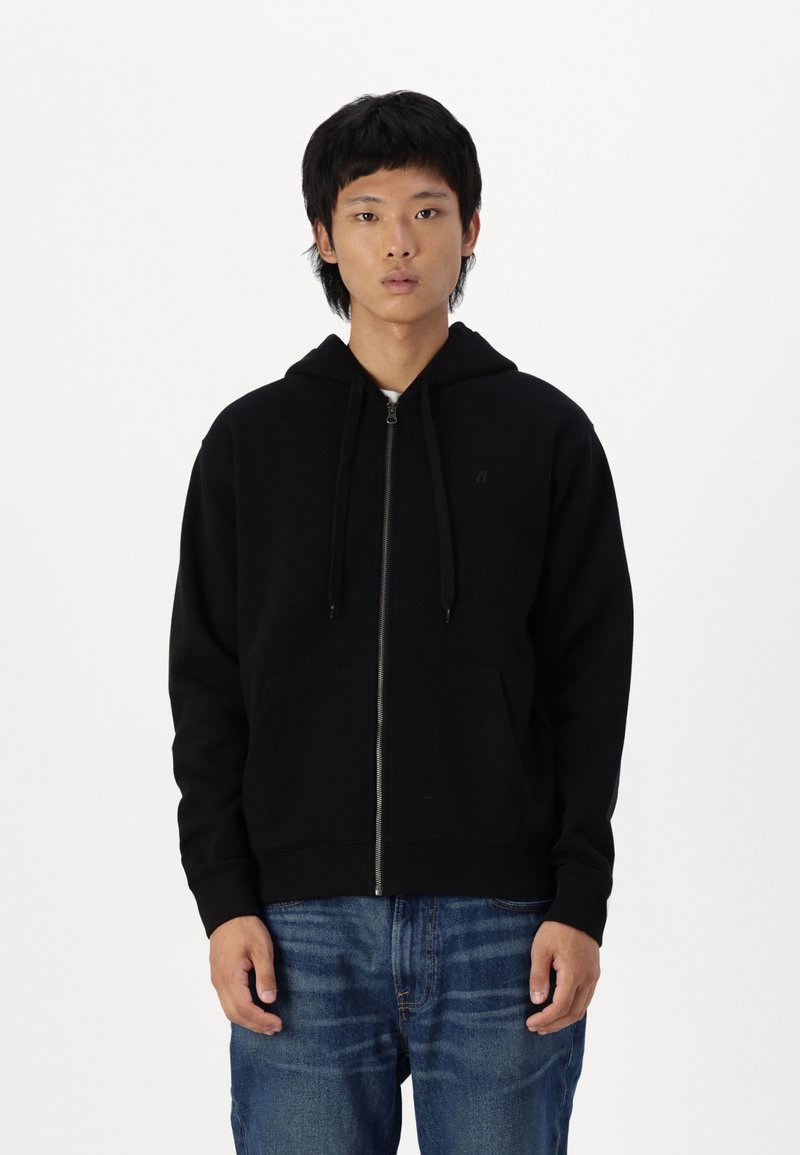 Zwarte rits hoodie met een voorkant zak, verstelbare koordjes en geribde manchetten, gemaakt van een zachte, gestructureerde stof. Draagt met blauwe jeans.