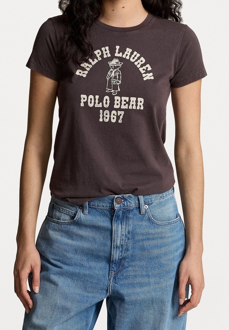 Kvinna som bär en mörkbrun Ralph Lauren-t-shirt med en Polo Bear-grafik och ljusblå högmidjade jeans.