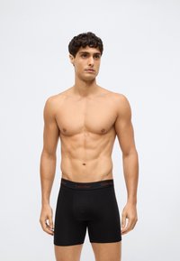 Fit man met krullend haar, gekleed in zwarte Calvin Klein boxershorts, staand tegen een effen witte achtergrond met een neutrale uitdrukking.