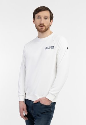 Homme aux cheveux bruns et à la barbe portant un sweat-shirt blanc avec un texte sur la poitrine et un jean bleu foncé, debout devant un fond blanc.