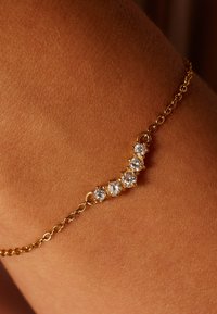 Gouden kettingarmband met vijf ronde heldere stenen in een gebogen opstelling. De armband ligt op een gebruinde huid.