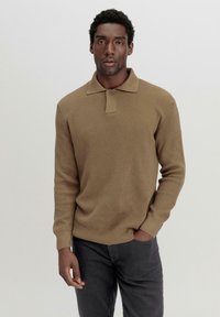 Braunes, geripptes Strick-Poloshirt mit Kragen und Knopfverschluss, kombiniert mit dunkelgrauen Hosen. Der Stoff wirkt weich und strukturiert.