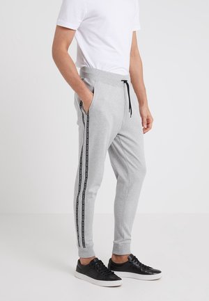 Mann trägt hellgraue Jogginghose mit schwarzen Seitenstreifen, weißes T-Shirt und schwarze Sneakers mit weißen Sohlen, steht vor weißem Hintergrund.