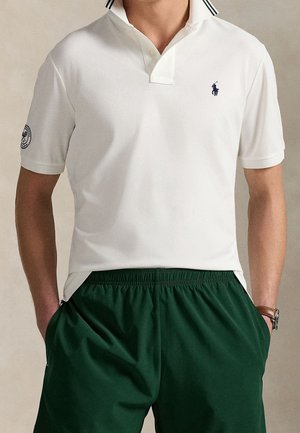 Man draagt een wit poloshirt met korte mouwen en een zwart logo op de borst en groene korte broek met elastische taille, staand met handen in de zakken.