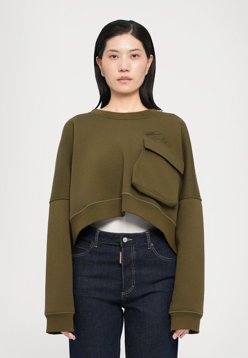 Felpa corta verde oliva con maniche lunghe, tasca frontale e dettagli di cucitura in contrasto. Indossata sopra jeans blu scuri.