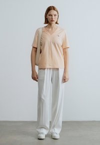 Ανοιχτό ροδακινί μπλουζάκι με v λαιμόκοψη σε συνδυασμό με λευκό παντελόνι φαρδιάς γραμμής. Λείο ύφασμα, μινιμαλιστικός σχεδιασμός, με διακριτικό λογότυπο στο μπλουζάκι.