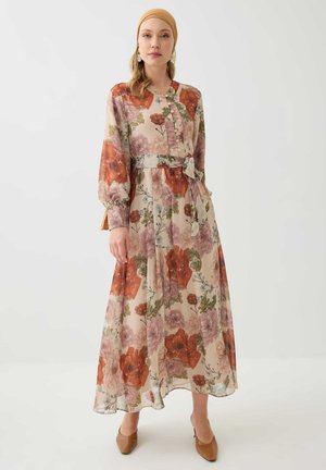 Mujer que lleva un vestido largo estampado con flores naranjas, rosas y verdes, combinado con una diadema beige y zapatos marrones, de pie frente a un fondo blanco.