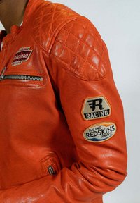 Veste de course en cuir rouge avec épaules matelassées, fermeture éclair argentée et écussons indiquant "RACING" et "REDSKINS SERVICE."