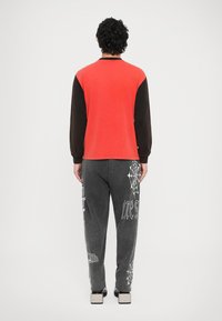 Crvena i crna majica dugih rukava s okruglim izrezom; sive trenirka hlače s bijelim motivima paukove mreže i tekstualnim detaljima na nozi.