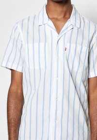 Camisa de manga corta con botones en tela blanca con rayas verticales azul claro. Presenta dos bolsillos en el pecho y un diseño de cuello tipo campamento.