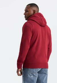Sudadera roja con capucha, fabricada en un tejido texturizado. Combinada con unos jeans de mezclilla claros, que muestran puños y dobladillo acanalados. Diseño simple, sin patrones.
