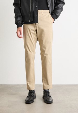Pantaloni in cotone beige con taglio dritto, dotati di tasche frontali e cintura con bottone. Abbinati a scarpe in pelle nere.