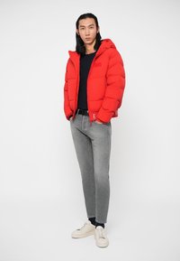 Moški v rdeči puhasti jakni, črni majici, sivih kavbojkah in belih supergah stoji z rokami v žepih ob beli ozadju.