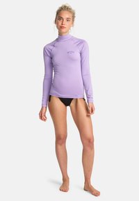 Rash guard lungo a maniche viola realizzato in materiale elastico, con un colletto alto e un piccolo logo sul petto. Indossato con pantaloni da bagno neri.