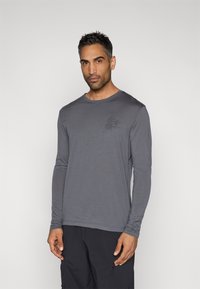 Mann med kort mørkt hår iført en langermet grå skjorte med et lite blomsterdesign på brystet og svarte bukser, stående mot en ensfarget bakgrunn.