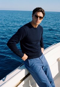 Homme portant des lunettes de soleil, un pull bleu marine et un pantalon bleu, appuyé sur une rambarde blanche de bateau avec un océan calme en arrière-plan lors d'une journée ensoleillée.