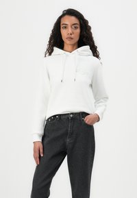 Femme aux cheveux bouclés portant un sweat à capuche blanc et un jean noir, debout avec une main dans la poche, sur fond blanc.