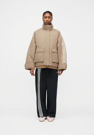 Ženska stoji in nosi bež puhasto jakno z belimi črtami na rokavih, črne hlače z belimi črtami in bledo roza čevlje na belem ozadju.