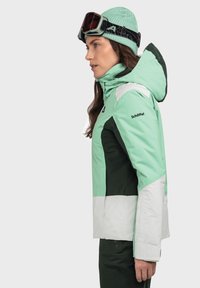 Skijacke in Mintgrün, Dunkelgrün und Weiß mit Kapuze. Verfügt über Reißverschlusstaschen und weichen Stoff. Getragen mit einer passenden Strickmütze und einer Skibrille.