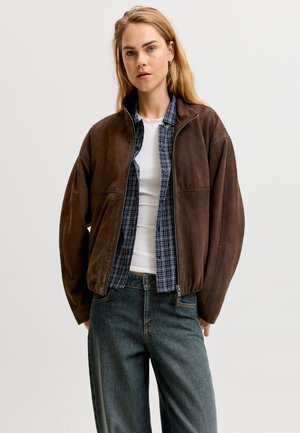 Femme aux longs cheveux blonds porte une veste marron zippée par-dessus une chemise bleue à carreaux et un haut blanc avec un jean foncé sur un fond uni.