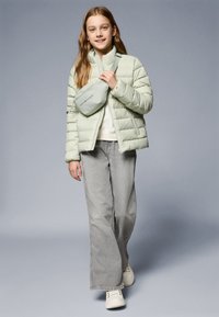 Casaco puff verde-claro, calças jeans largas cinzentas e ténis brancos; inclui uma pochete correspondente com detalhe do logótipo. Textura suave.