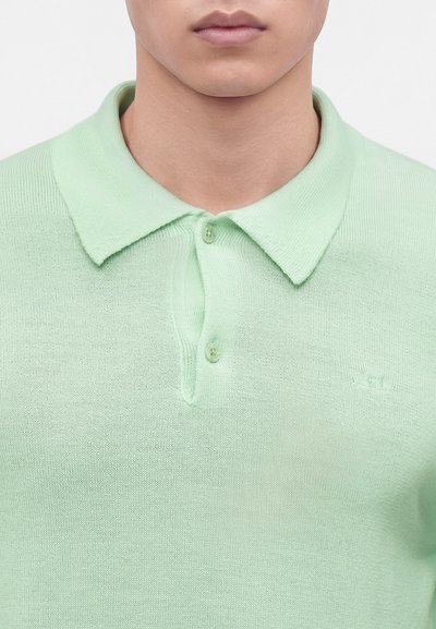 Gros plan d'une personne portant un polo tricoté vert clair avec deux boutons fermés et un col plié.