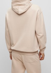 Sudadera beige de tela suave con capucha ajustable y puños de canalé. Combinada con pantalones jogger a juego que cuentan con un bolsillo trasero.