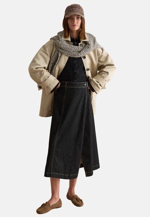 Femme portant une veste beige, une robe en denim foncé à boutons, une écharpe tricotée, une casquette marron et des mocassins marron à glands, debout les mains dans les poches.