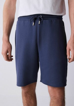 Mann trägt marineblaue Shorts mit Kordelzug und ein weißes T-Shirt, zeigt die Länge bis zur Mitte des Oberschenkels und Seitentaschen vor neutralem Hintergrund.