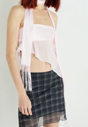 Frau trägt ein blassrosa asymmetrisches trägerloses Oberteil mit Schal und einen schwarzen transparenten karierten Rock vor weißem Hintergrund.