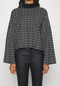 Black and white patterned sweater with a high neck and bell sleeves, made of textured fabric. Paired with black fitted pants.