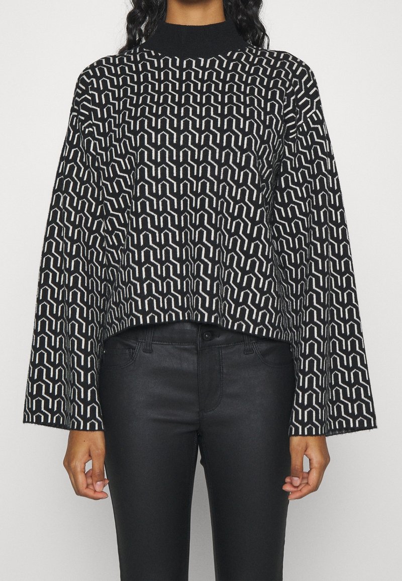 Black and white patterned sweater with a high neck and bell sleeves, made of textured fabric. Paired with black fitted pants.