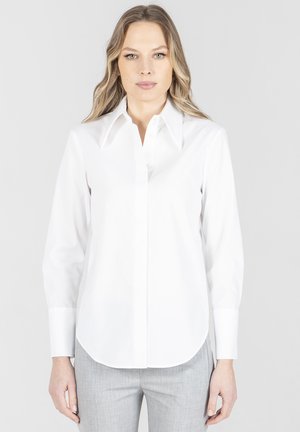 Kobieta o długich blond włosach, ubrana w białą koszulę na guziki i jasnoszare spodnie, stojąca na tle jednolitego jasnego tła.