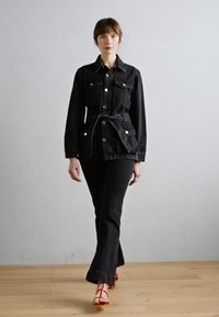 Black denim jacket with front pockets and tied belt; paired with flared black denim pants; model wearing red sandals; neutral background.