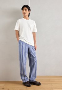 Λευκό κοντομάνικο μπλουζάκι με τσέπη, συνδυασμένο με μπλε και λευκές ριγέ χαλαρές παντελόνες και σκούρες καφέ loafers. Λείο ύφασμα.