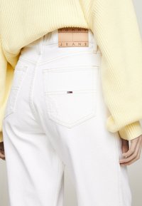 Jean de mezclilla blanca con un corte relajado, que presenta bolsillos traseros y una etiqueta de cuero con la inscripción "TOMMY JEANS". Llevados con un suéter amarillo claro.