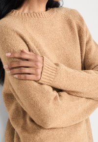 Kobieta w miękkim, wełnianym swetrze w kolorze camel z prążkowanymi mankietami, z założonymi rękami, ukazująca teksturę i dopasowanie w górnej części ciała.