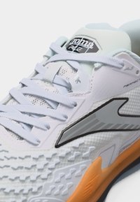 Gros plan sur une chaussure de sport blanche avec semelle texturée, logo gris sur le côté, lacets blancs et semelle extérieure en caoutchouc orange.